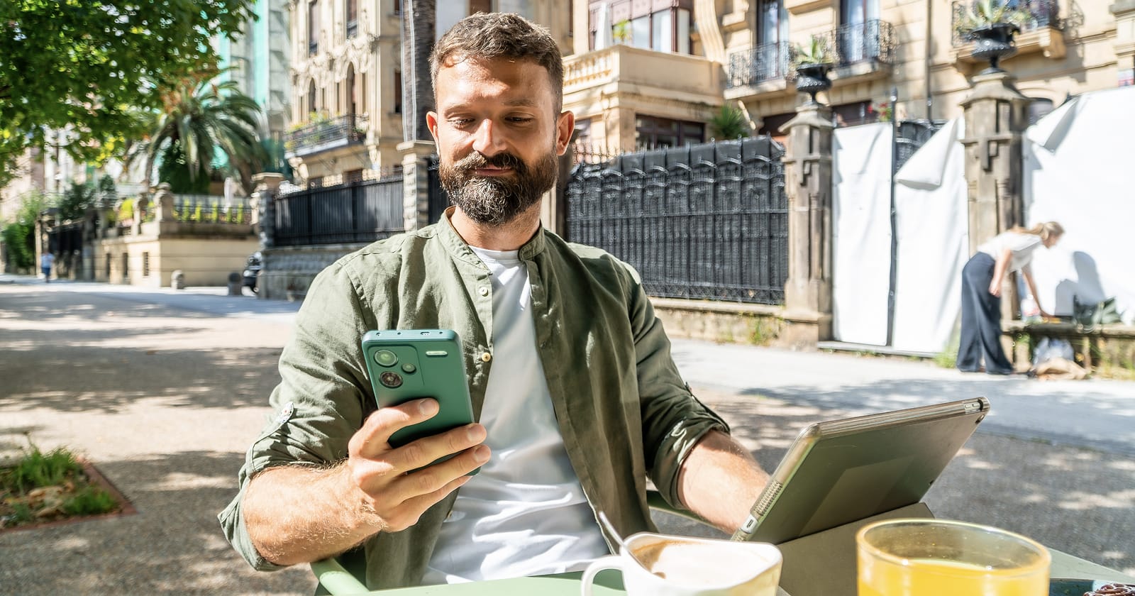 Man using a smartphone at an outdoor café table with a tablet beside him.
